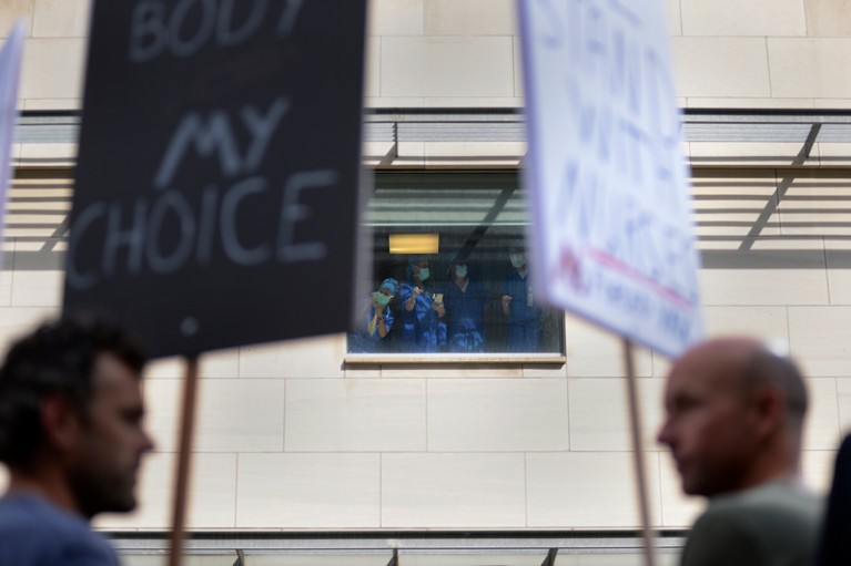 Hospital workers wearing PPE watch a protest through a hospital window