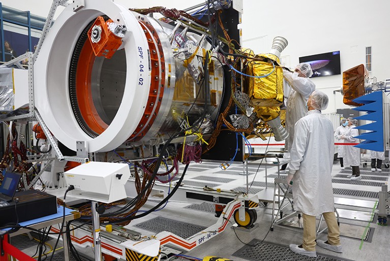 Two people in white coats inspect part of a satellite in a spacecraft assembly facility