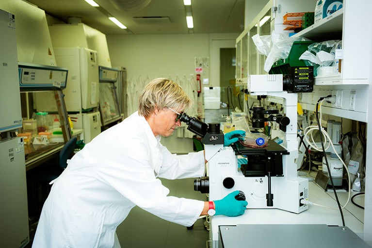 A person wearing a lab coat and green gloves is standing in a laboratory and looking into a microscope