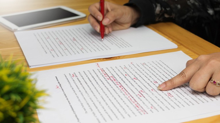 A person reviews and edits a paper using a red pen, with a tablet in the background.