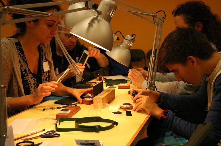 A group of people at a cluttered workbench use tools and smartphones to take macro photographs of mosquitos