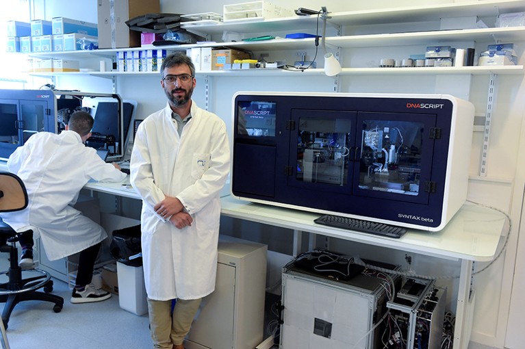 Man wearing white coat stands in laboratory next to benchtop DNA printer.