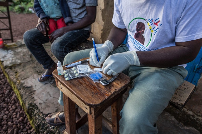 An individual examines a rapid diagnostic test set up on a stool outside a rural clinic; a parent and child wait to their right.