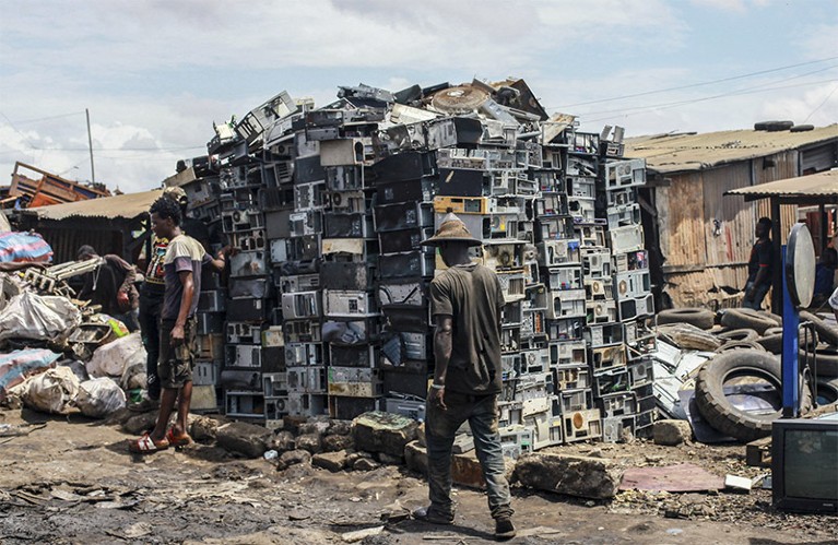 A man walks past a large stacked pile of electronics at Agbogbloshie e-waste dump in Ghana