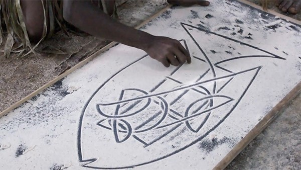 A person off-frame is in the middle of drawing a complex pattern resembling a large fish onto a sand-covered surface with their finger.