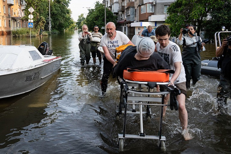 A woman wearing a woolly hat is pushed on a stretcher through flooded ground by two men. People around them are taking photos.