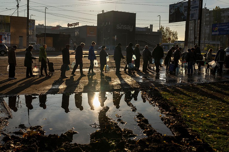 A long queue of adults at sunset, holding large plastic bottles. Many of them are reflected in a large puddle on the ground.