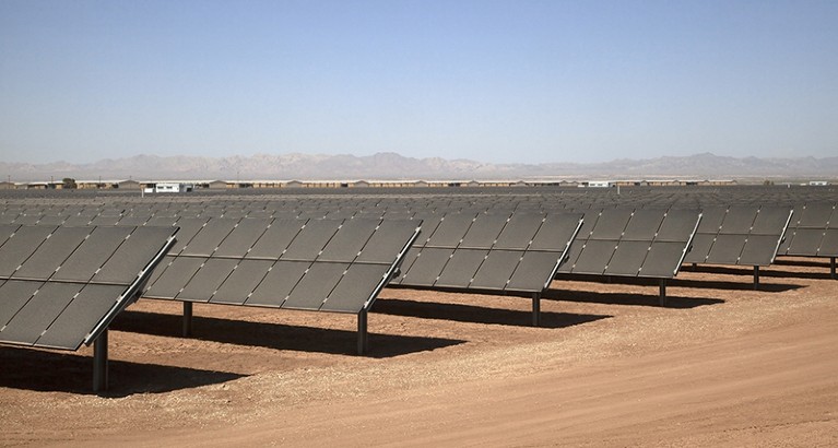 Arid, sandy ground with multiple rows of rectangular solar panels on it. Far off in the background are houses and mountains.