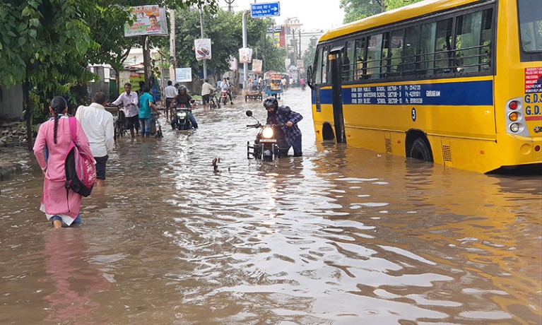 Water-looged street is seen with yellow bus on the right and people walking through water on the left