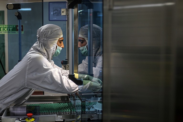 Person wearing protective clothing, mask, and gloves, works with vaccine vials on production line