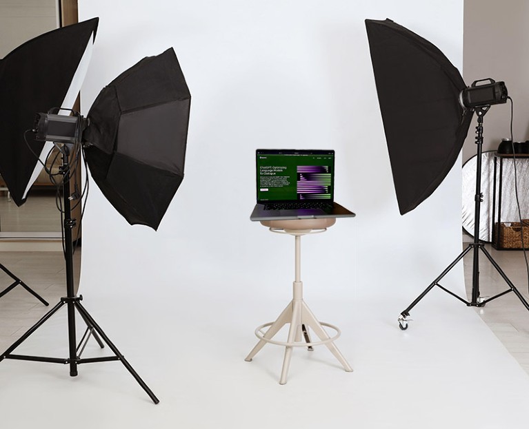 A composite artwork of a laptop displaying the OpenAI ChatGPT website on a stool surrounded by professional lighting equipment in a photo studio.