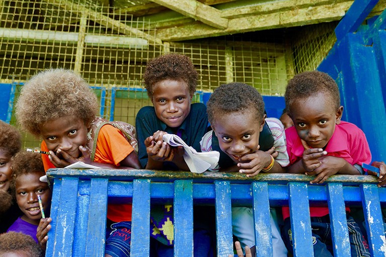 Group of Black children lean on a blue wooden fence, smiling to camera.