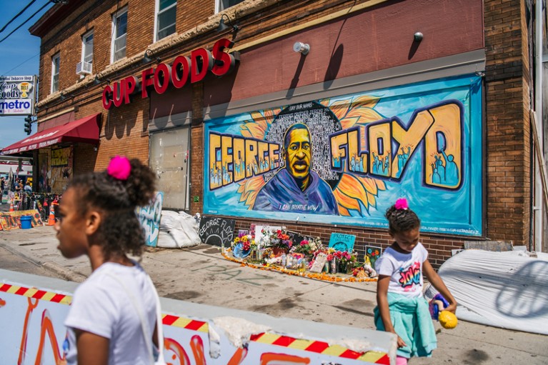 Two young girls walk in front of a mural for George Floyd that has flowers placed underneath