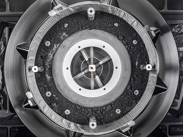 A top-down view of a round, gleaming metal container with the lid removed, revealing dark dust and rocks inside.