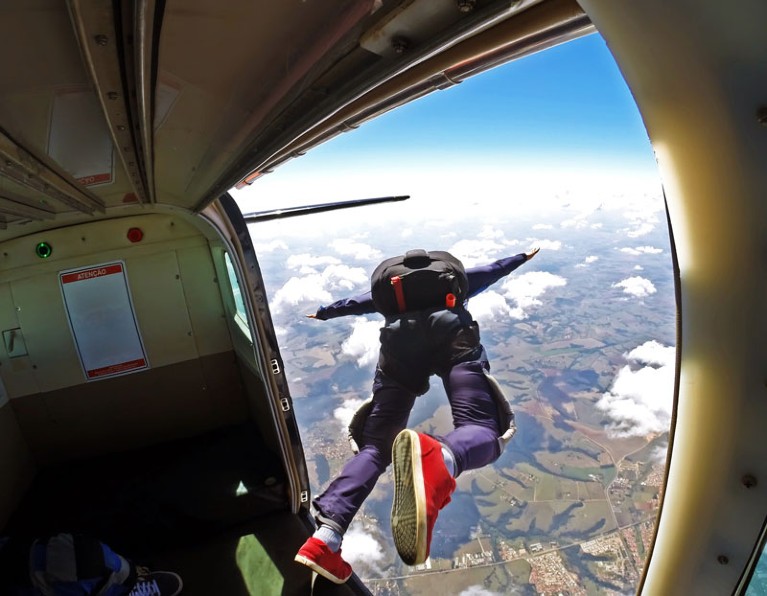 View from inside a plane flying over clouds of a skydiver wearing a parachute and red trainers jumping out of the plane