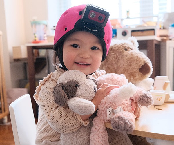 Photo of a young toddler in a light pink onesie sitting on a chair and smiling at the camera while clutching several stuffed animals. The child is wearing a bright pink helmet fitted with a head-mounted camera.