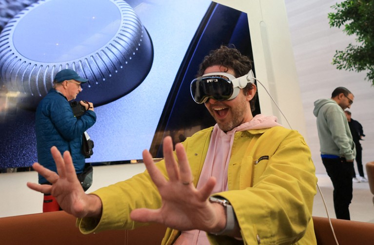 A man wearing a yellow jacket and an Apple Vision Pro headset examines his hands while smiling