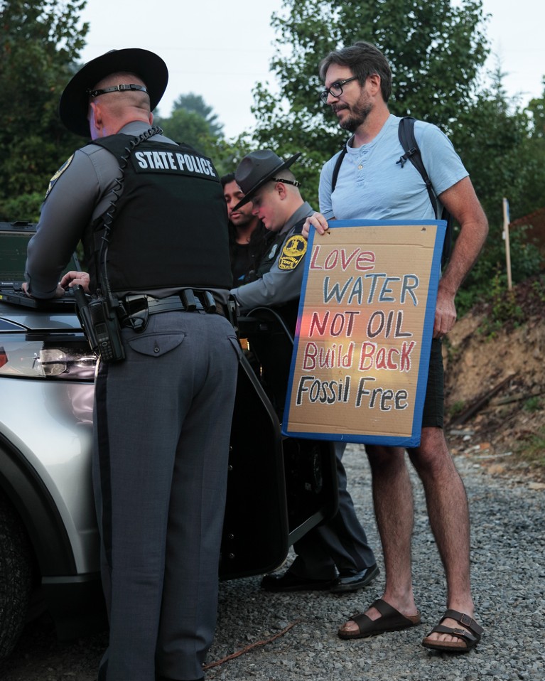 Peter Kalmus holding a protest board looks on as Virginia State Police take down his information