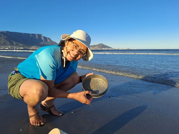A smiling young woman wearing shorts and sandals with a blue t-shirt and a cream sunhat crouches on a sandy beach while holding a stainless steel sieve. It is a sunny day with blue skies, and the sea is breaking gently behind her.