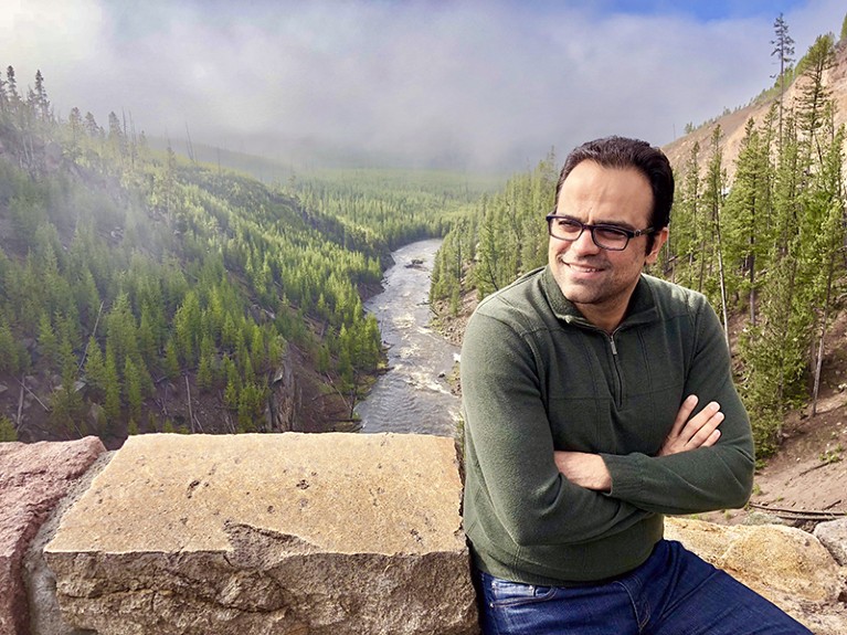 Morteza Mahmoudi sitting on a rock wall above a stream and forest.