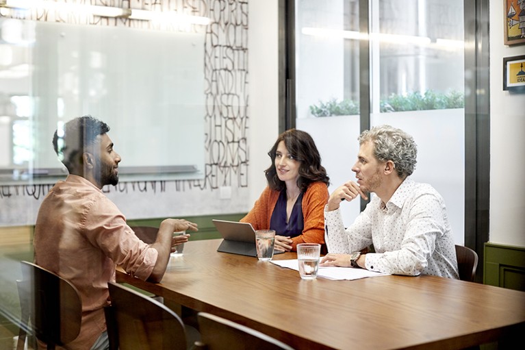 Three people sit at a table, talking in an office conference room.