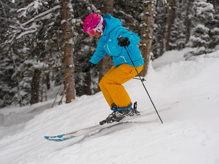 A woman in pink helmet, blue coat and yellow trousers skiing downhill with trees in the background.