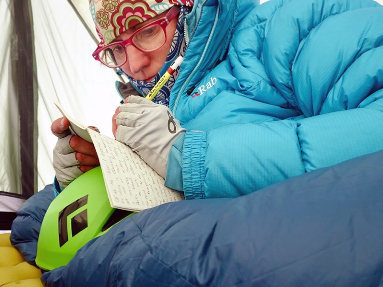 A woman in a blue coat and gloves writing in a notebook while wrapped in a sleeping bag.