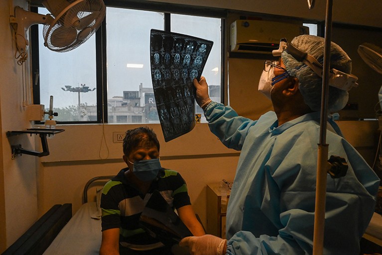 Person seated in room below window, wearing a face mask. To the right, person standing wearing PPE looking at MRI