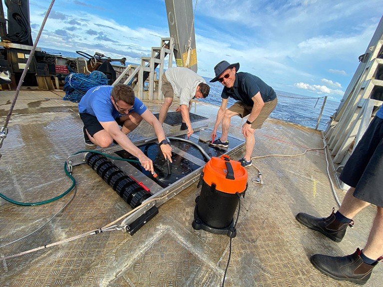 Three people use a vacuum tool on a metallic sledge on board a ship.