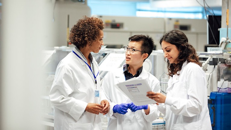 A group of three female technicians discuss work in laboratory while wearing white lab coats.