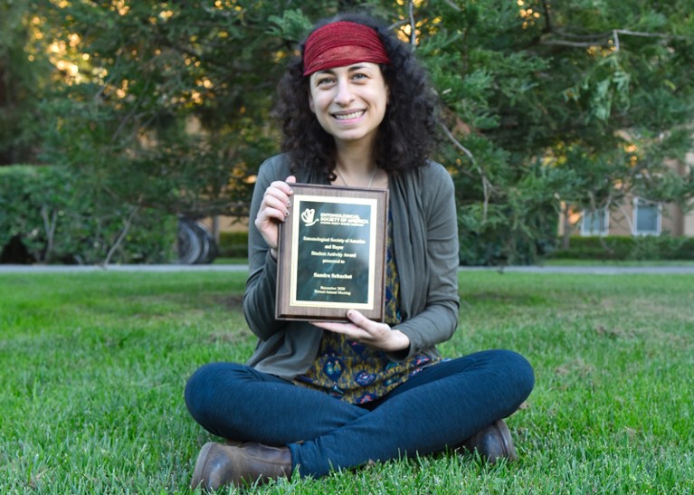 Sandra R. Schachat sits on the grass holding an award