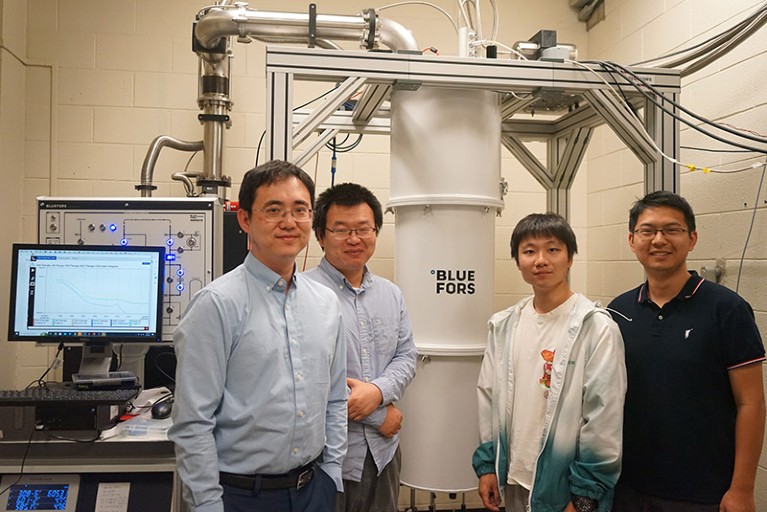 Four people standing next to a computer and a cryogenic measuring system.