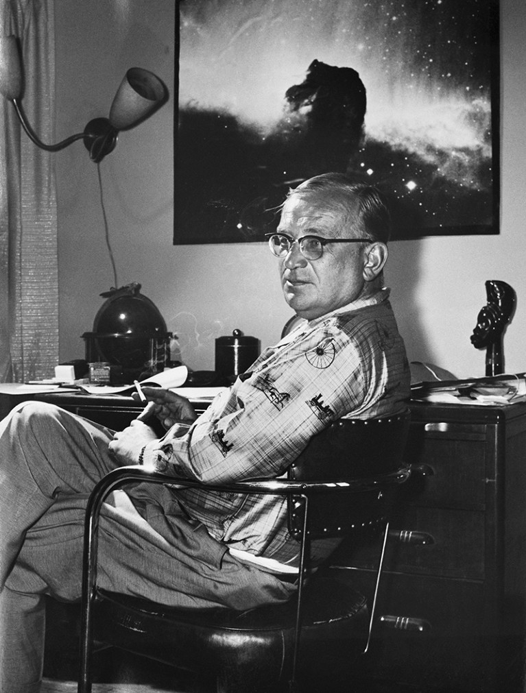 George Gamow sitting in a chair at a desk in front of a celestial photograph hanging on the wall