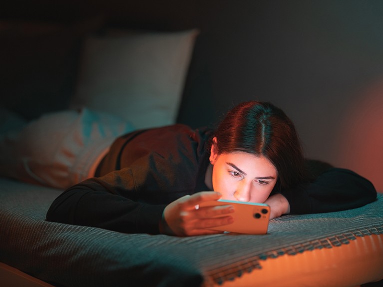 A teenage girl lies on the bed in her room lightened with orange and teal neon lights and watches a movie on her mobile phone.