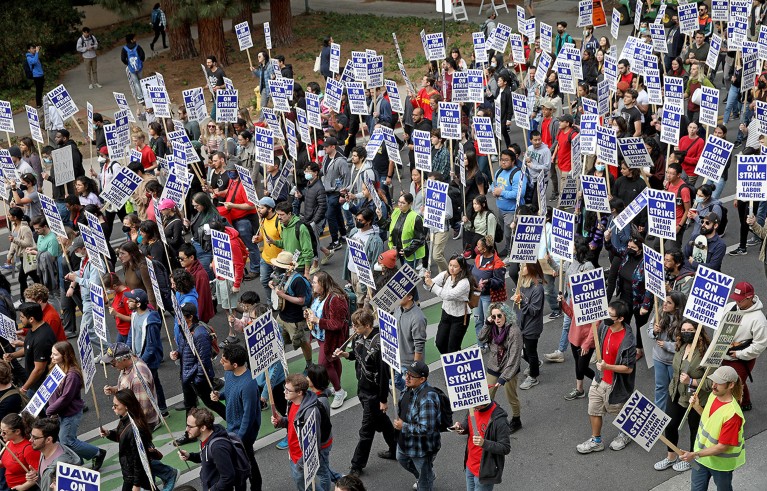 A crowd of people, many wearing red t-shirts or high-visibility jackets, holding blue and white placards.