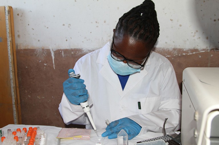 Person sitting at table, wearing white coat, blue gloves and blue face mask, holding pipette in one hand