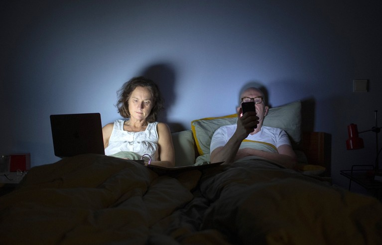 A woman and a man sit in bed in a dark bedroom, distracted by a laptop computer and a smartphone respectively.
