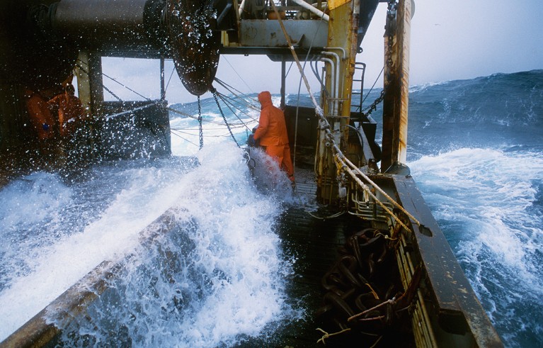 A fishing trawler hauls back its net on the Bering Sea with a fisherman in an orange vest navigating the high sea.
