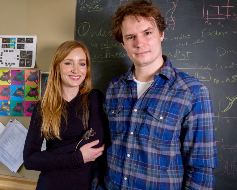 Mackenzie Mathis holding a mouse posing for a portrait with Alexander Mathis standing in front of a blackboard with chalk writing on it