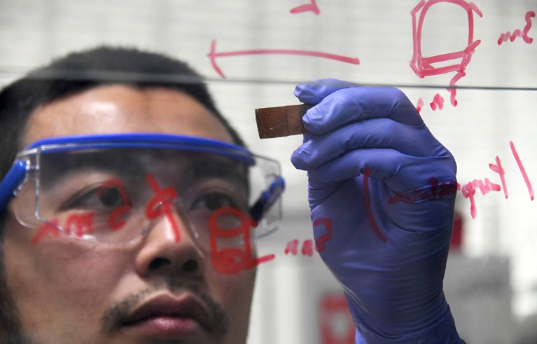 Viewed through a window covered in red handwritten notes, a man wearing safety goggles holds a piece of repaired broken resin glass.