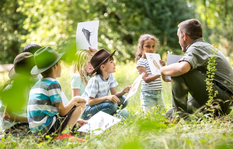 Six school children doing nature studies with their teacher, hold pictures of animals they have drawn.