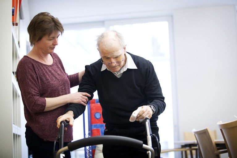 An older man walks using a zimmer frame while a woman supports him by the arm.