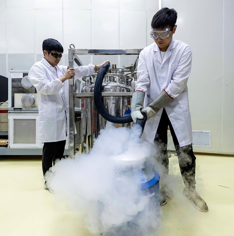 Dos personas con batas blancas de laboratorio, guantes y gafas protectoras se encuentran a ambos lados de una gran tina que vierte nitrógeno líquido.