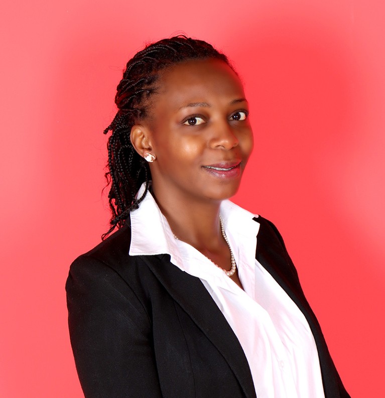Winnifred Kansiime wearing a black jacket and white shirt, looking at camera, photographed against red background