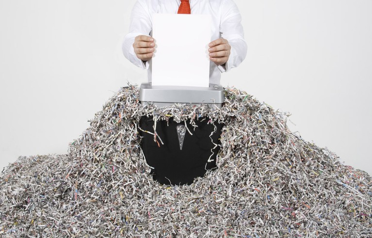 A person stands amongst a large mound of shredded paper documents while inserting a white piece of paper into a shredder.