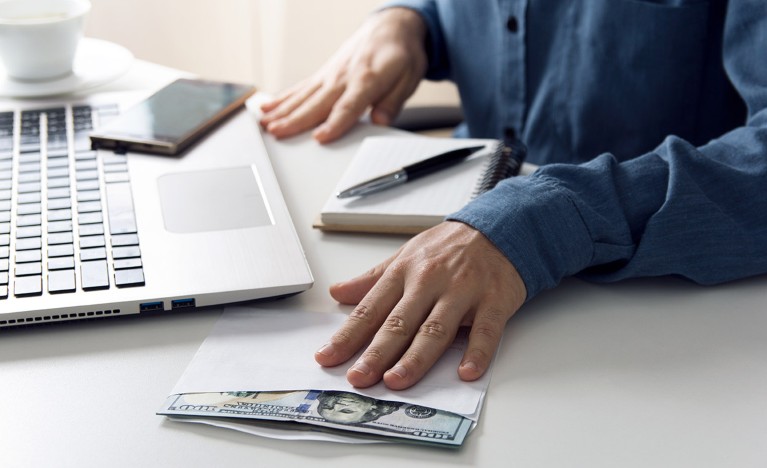 Close up of a desk and laptop with a man's hand resting on an envelope filled with US dollar bills.