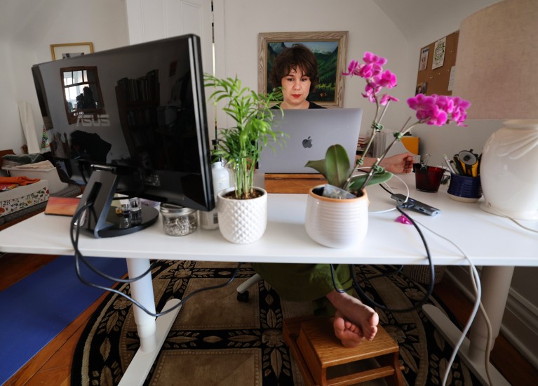 A woman works on the laptop at her desk in her home. A monitor and two plants are also seen on the desk