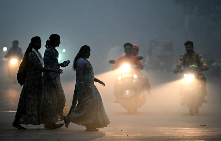 Tres mujeres caminan por una calle cubierta de smog en Chennai, temprano en la mañana, mientras algunas motocicletas pasan al fondo.