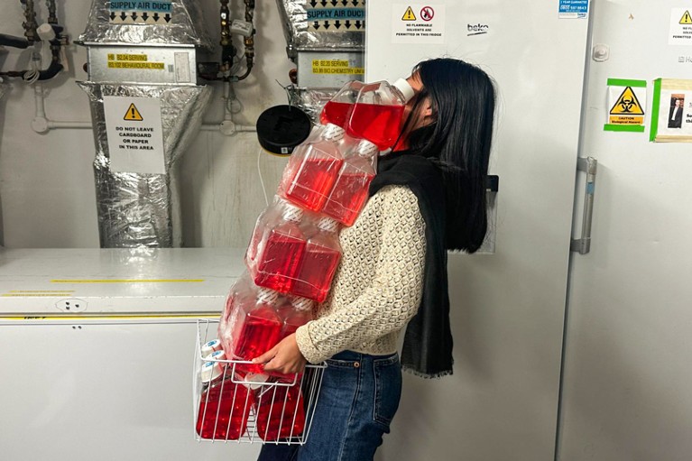 Un trabajador de laboratorio sostiene varios frascos de medios de cultivo de glóbulos rojos frente a los congeladores y conductos del laboratorio.