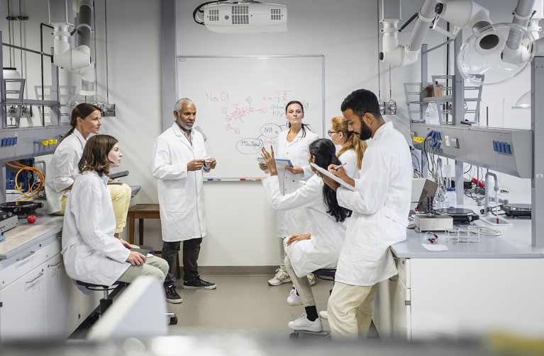 A group of seven scientists in lab coats are meeting in a lab.